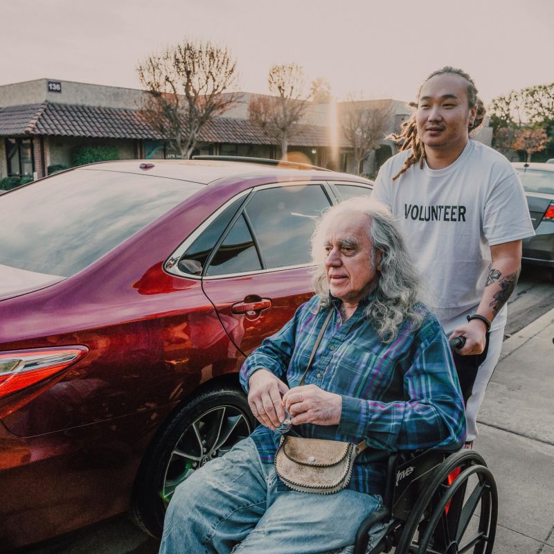 Caregiver helping a man in wheelchair from his car.